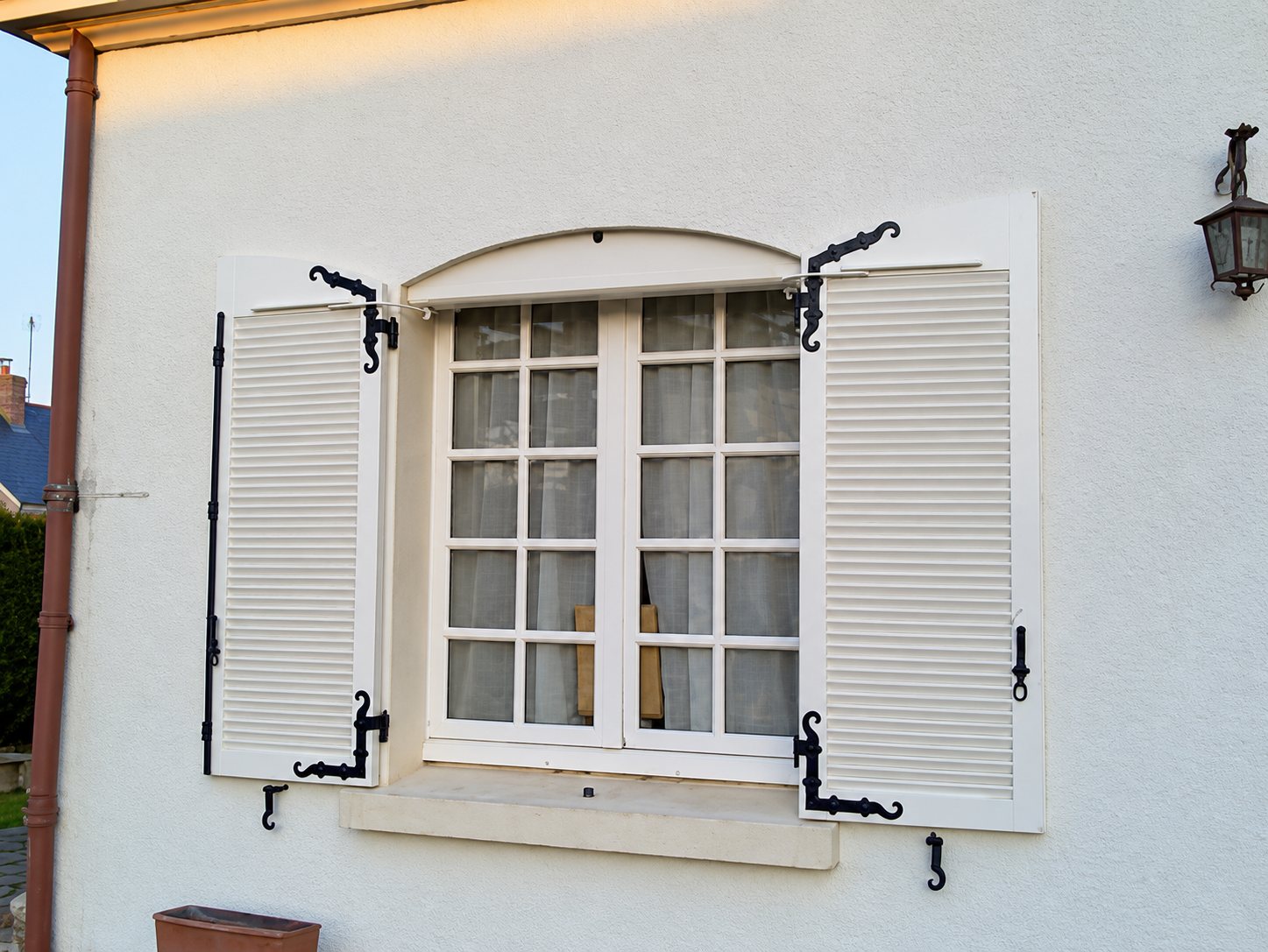 Fenêtre à petits carreaux encadrée de deux volets persiennes blancs ouverts, avec ferrures noires, sur un mur blanc éclairé par le soleil couchant.
