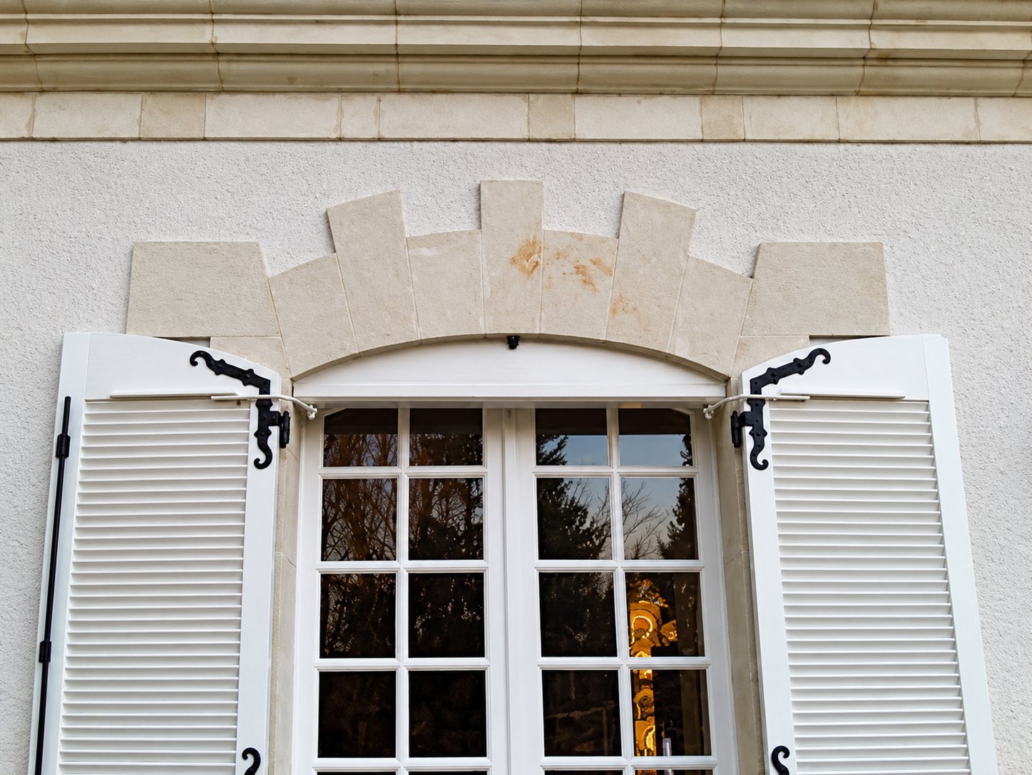 Fenêtre à petits carreaux encadrée de deux volets persiennés blancs avec ferrures noires, surmontée d'un linteau en pierre claire sur une façade enduite.