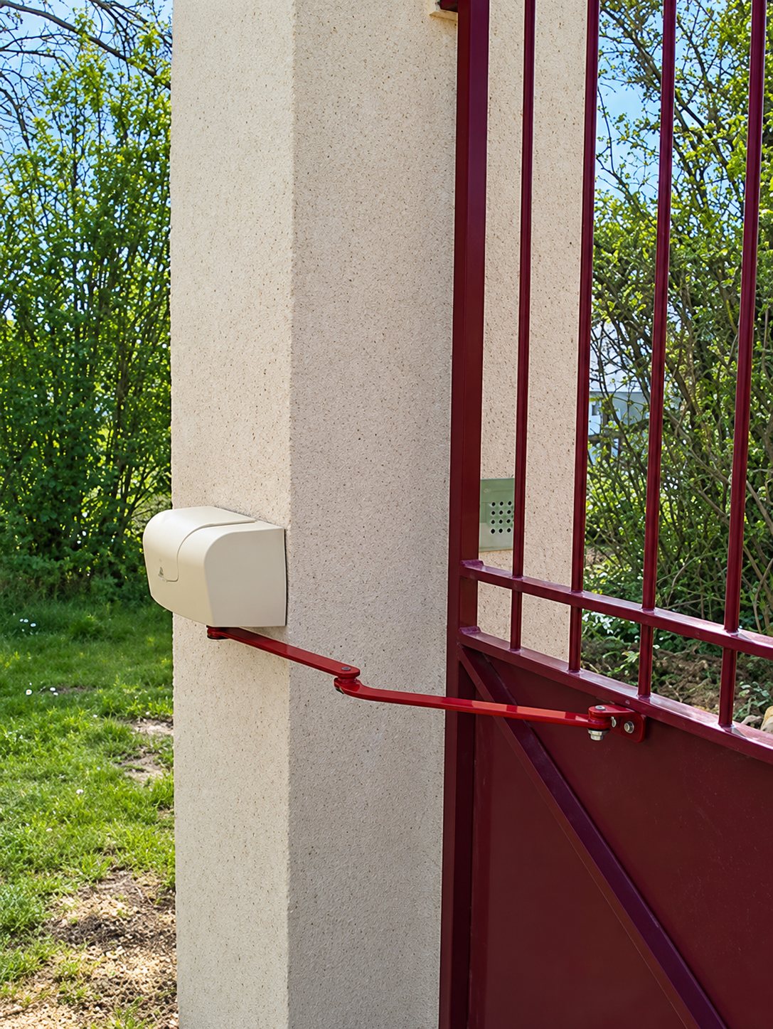Un moteur de portail beige à bras rouge est fixé sur un pilier en crépi clair, à côté d'un portail métallique bordeaux dans un jardin.