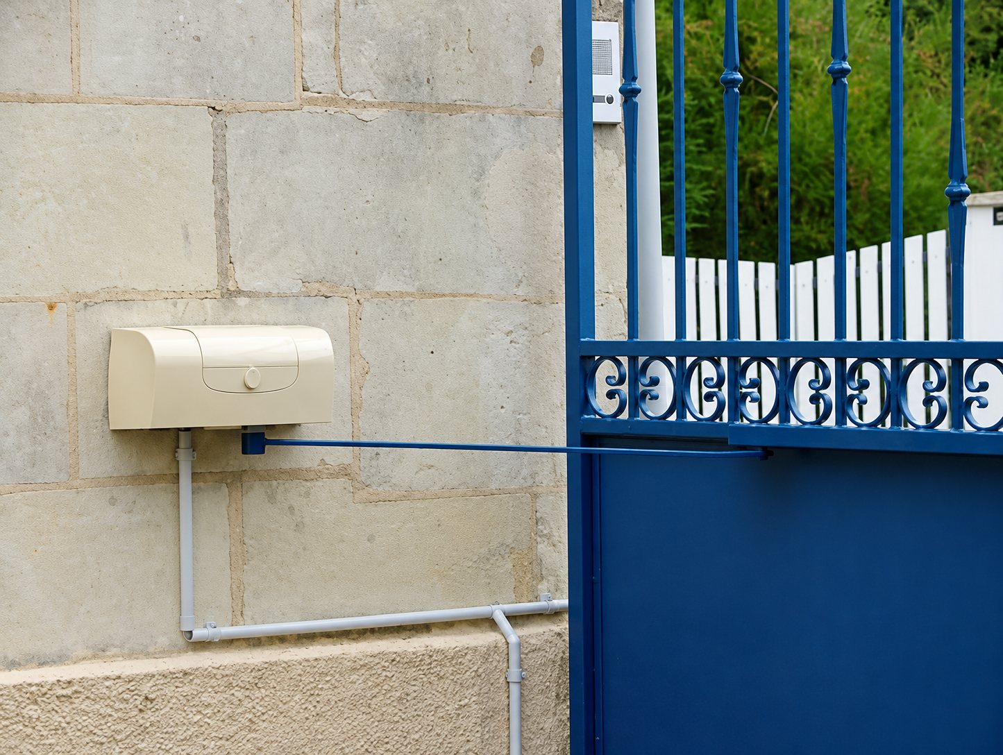 Un portail bleu en fer forgé est équipé d'un boîtier de motorisation beige fixé sur un mur en pierre claire, relié par un bras et des gaines grises.