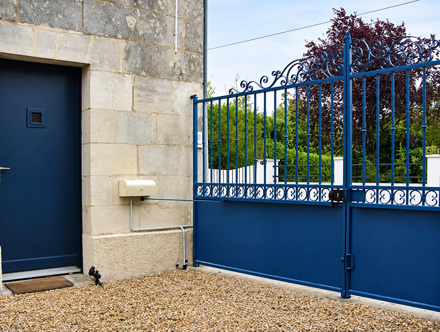 Un portail bleu en fer forgé aux motifs ornementaux se tient à côté d'une porte bleue dans un mur en pierre claire, avec des arbres en arrière-plan.