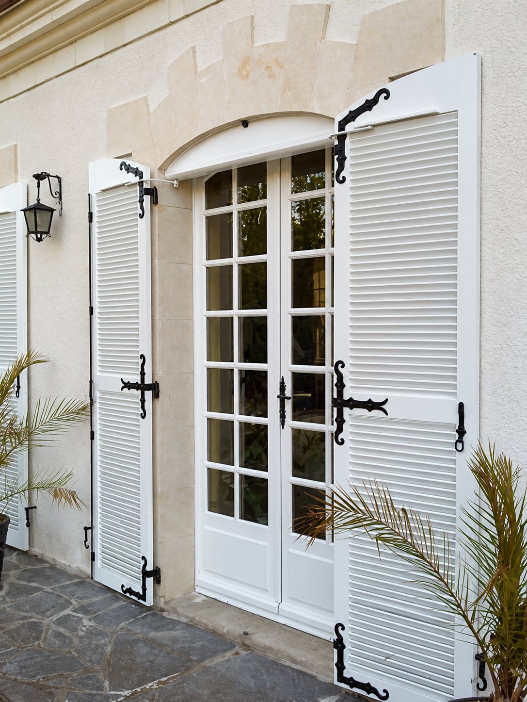 Une porte-fenêtre à petits carreaux encadrée de deux volets persiennes blancs ouverts, avec ferrures noires, sur une façade claire ornée d'une lanterne.